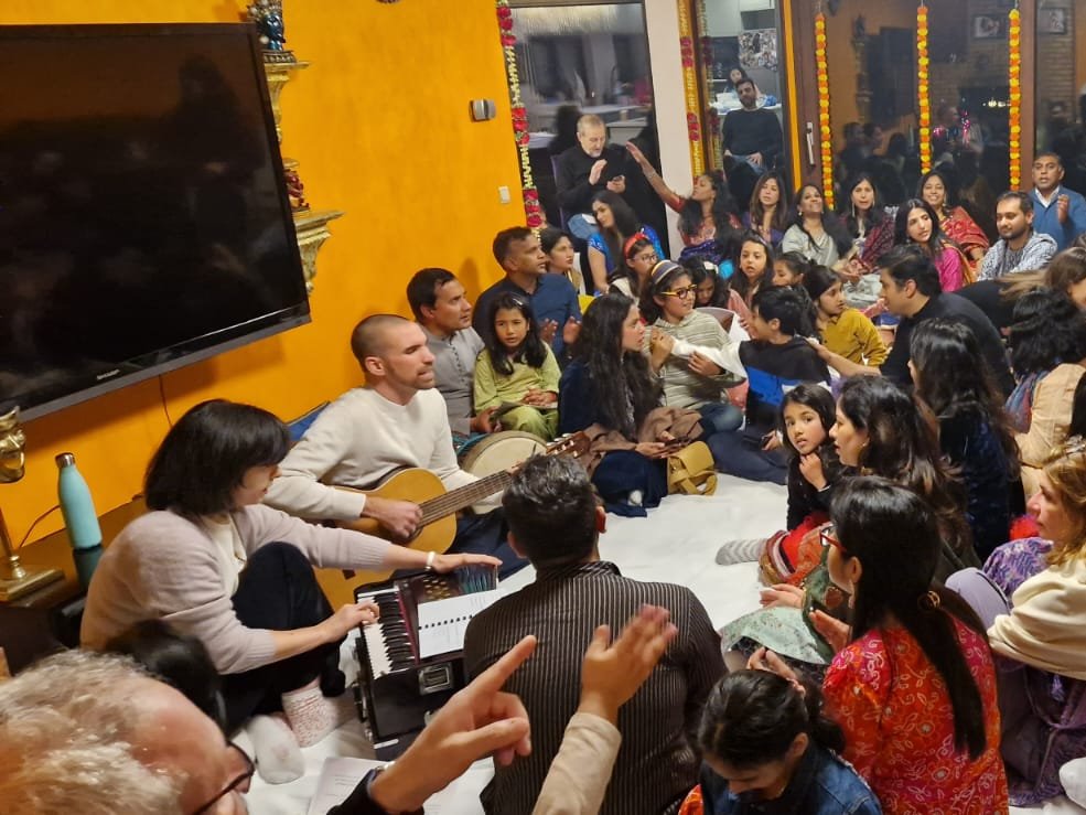 Jorn Ayan playing guitar with a harmonium player, leading a full room in a home kirtan circle